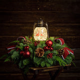 An arrangement of noble fir, incense cedar, and white pine with red ball clusters, pinecones, red berry clusters, red linen bows with white edges, and cardinals in a globe lantern lit up with a wood background.