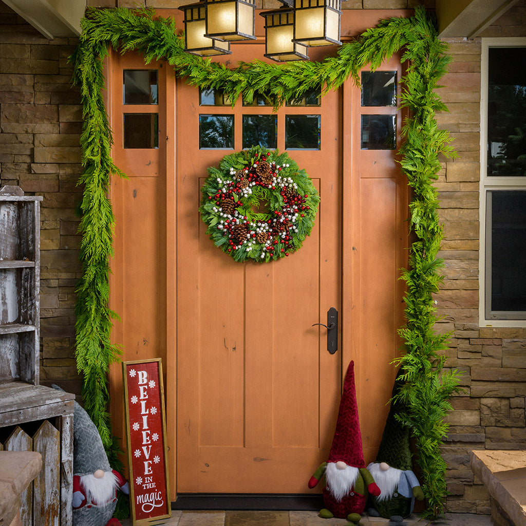 Framed wood sign engraved with "Believe in the Magic" in white while falling snowflakes surround the words on a front porch surrounded by the cascade christmas wreath, garland, and plush holiday gnomes. 