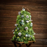 18" tree made of noble fir, glittery mint and silver ball clusters, silver berry clusters, frosted Austrian pinecones, small Chickadee birds, a bird’s nest, and a strand of warm white lights with a dark wood background.