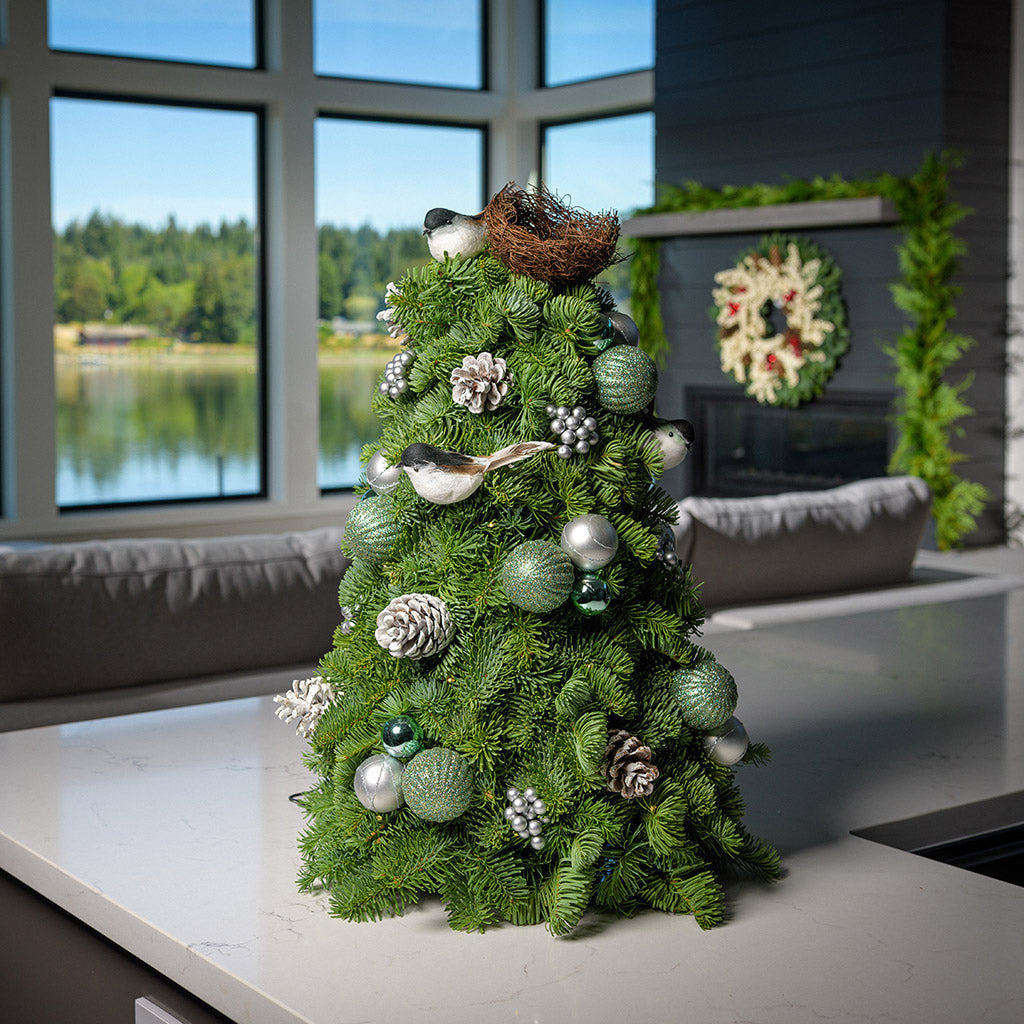18" tree made of noble fir, glittery mint and silver ball clusters, silver berry clusters, frosted Austrian pinecones, small Chickadee birds, a bird’s nest, and a strand of warm white lights on a counter.