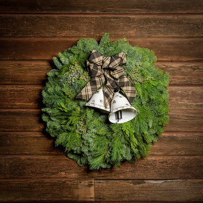 A wreath made of noble fir, incense cedar, juniper, and white pine with white metal bells, and a black and tan plaid linen bow on a dark wood background.