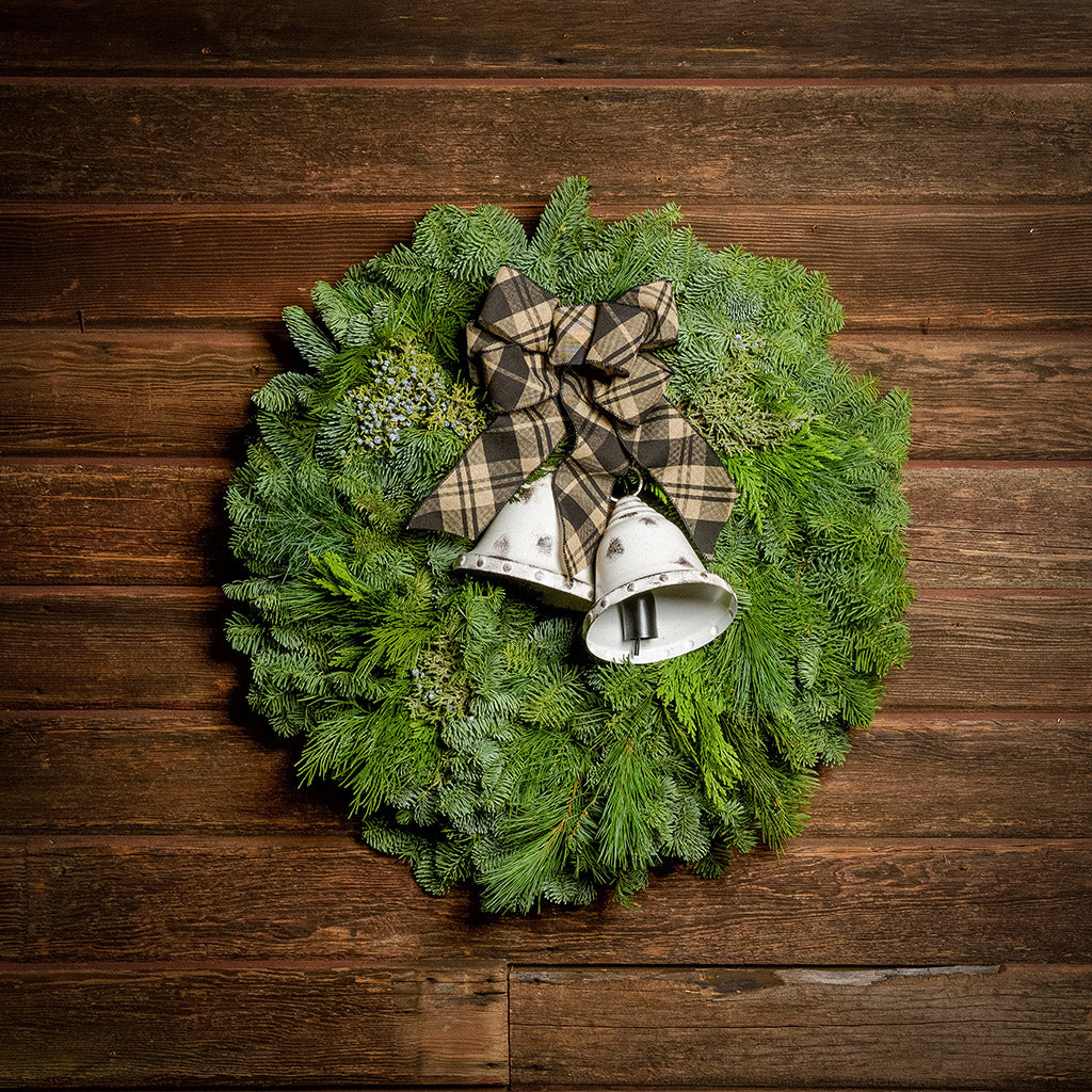 A wreath made of noble fir, incense cedar, juniper, and white pine with white metal bells, and a black and tan plaid linen bow on a dark wood background.