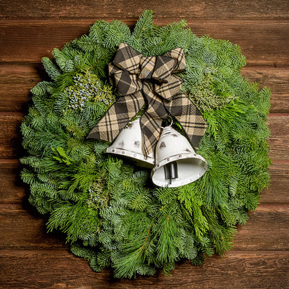 A wreath made of noble fir, incense cedar, juniper, and white pine with white metal bells, and a black and tan plaid linen bow on a dark wood background.