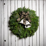 A wreath made of noble fir, incense cedar, juniper, and white pine with white metal bells, and a black and tan plaid linen bow on a white wood background.