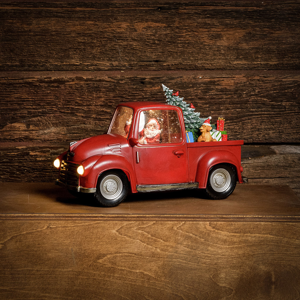 A Lighted Water Globe of Santa driving a truck with a tree, dog, and presents in the back with a dark wood background.