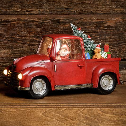 A Lighted Water Globe of Santa driving a truck with a tree, dog, and presents in the back with a dark wood background.