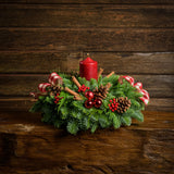 Centerpiece made of noble fir, white pine, incense cedar, red ball clusters, cinnamon-scented Australian pinecones, red and white plaid bows, real cinnamon sticks, red berry clusters, and a red pillar candle on a wooden table against a wood wall