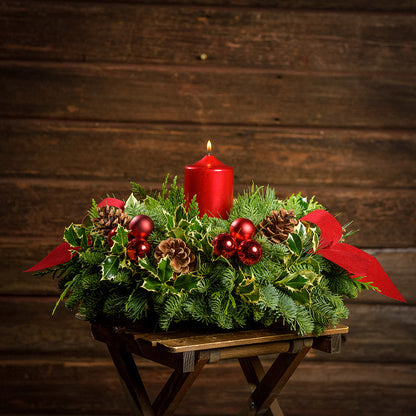 Christmas centerpiece with holly, pine cones, red ornaments and a red pillar candle with a dark wood background.