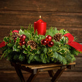 Christmas centerpiece with holly, pine cones, red ornaments and a red pillar candle with a dark wood background.
