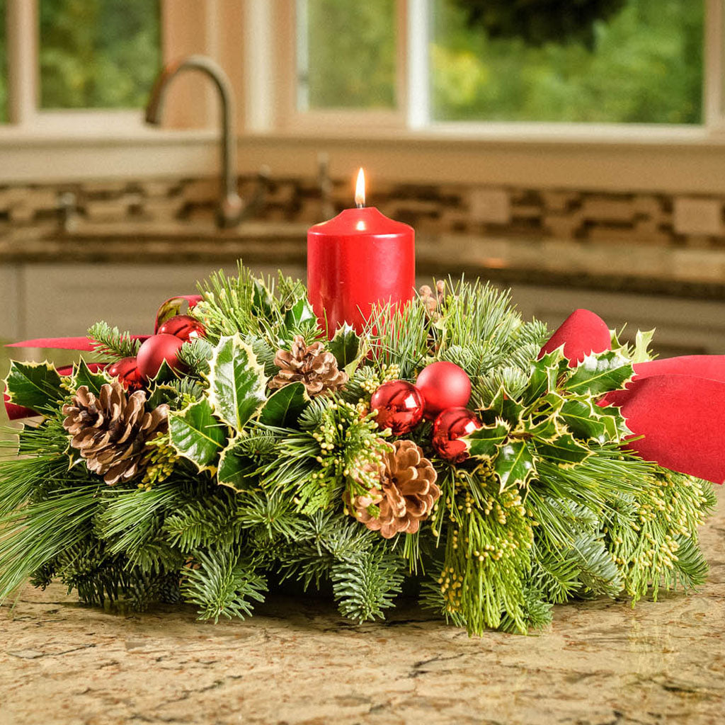 Christmas centerpiece with holly, pine cones, red ornaments and a red pillar candle displayed on a kitchen counter