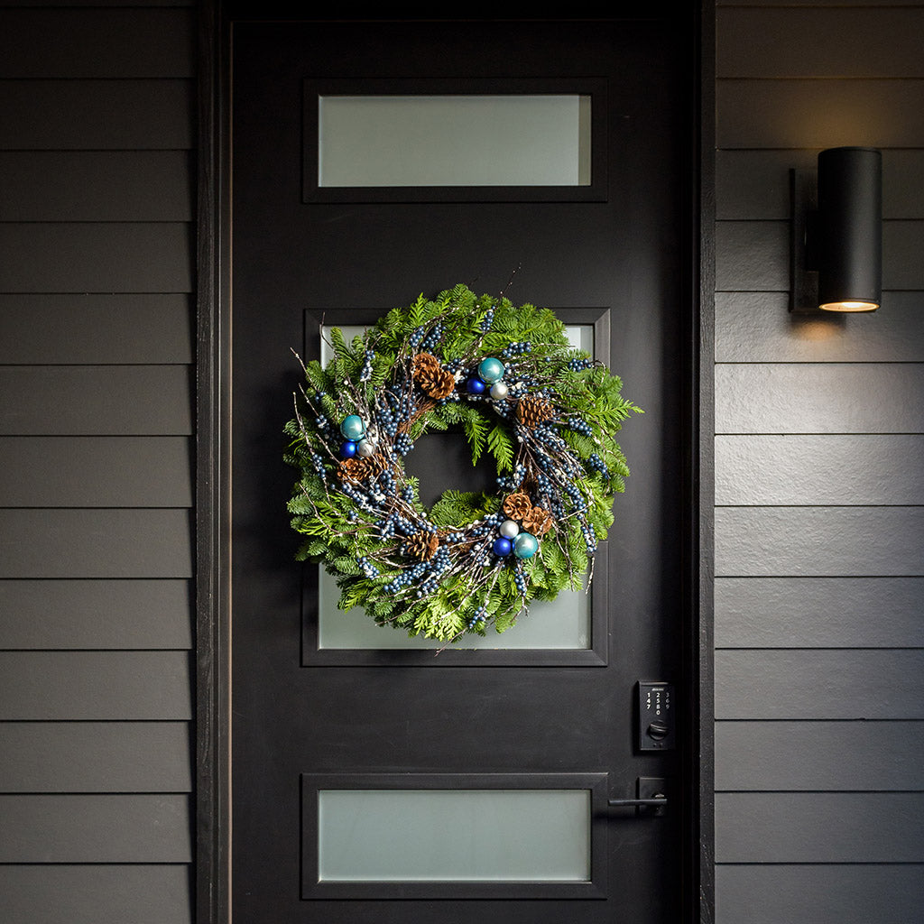 A holiday wreath of noble fir and western red cedar with a ring of faux metallic-blue berries and frosted branches, 3 blue and silver ball clusters, and 8 Australian cones on a black front door.