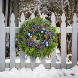 A holiday wreath of noble fir and western red cedar with a ring of faux metallic-blue berries and frosted branches, 3 blue and silver ball clusters, and 8 Australian cones on a white picket fence.