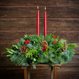 Centerpiece made of noble fir, pine, incense cedar and bay leaves with egg-sized ponderosa pine cones, red berry clusters, and 2 red 12" taper candles on a wooden bench