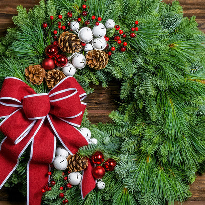 A holiday wreath made of noble fir and white pine with white jingle bells, 3 red berry clusters, 5 Australian pinecones, 2 red ball clusters, and a brushed red linen bow with white edging on a wood background.