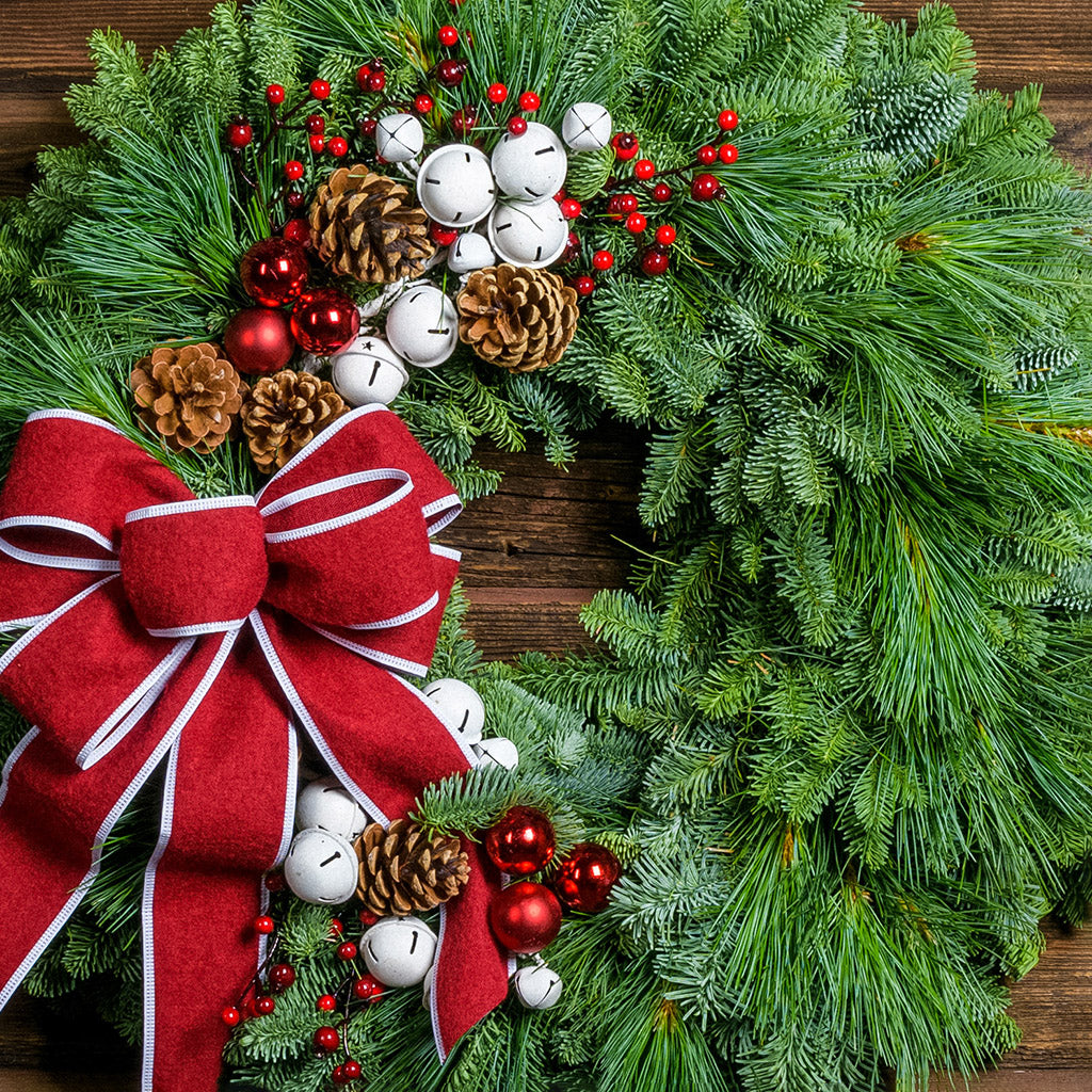 A holiday wreath made of noble fir and white pine with white jingle bells, 3 red berry clusters, 5 Australian pinecones, 2 red ball clusters, and a brushed red linen bow with white edging on a wood background.