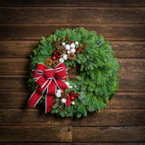 A holiday wreath made of noble fir and white pine with white jingle bells, 3 red berry clusters, 5 Australian pinecones, 2 red ball clusters, and a brushed red linen bow with white edging on a wood background.
