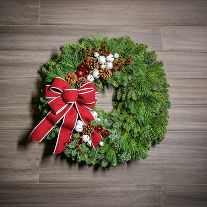 A holiday wreath made of noble fir and white pine with white jingle bells, 3 red berry clusters, 5 Australian pinecones, 2 red ball clusters, and a brushed red linen bow with white edging on a wood background.