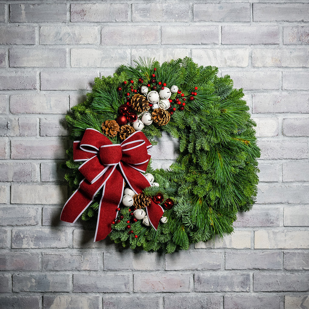 A holiday wreath made of noble fir and white pine with white jingle bells, 3 red berry clusters, 5 Australian pinecones, 2 red ball clusters, and a brushed red linen bow with white edging on a white brick background.