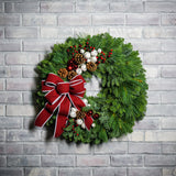 A holiday wreath made of noble fir and white pine with white jingle bells, 3 red berry clusters, 5 Australian pinecones, 2 red ball clusters, and a brushed red linen bow with white edging on a white brick background.