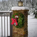 A holiday wreath made of noble fir and white pine with white jingle bells, 3 red berry clusters, 5 Australian pinecones, 2 red ball clusters, and a brushed red linen bow with white edging on a natural brick pillar.