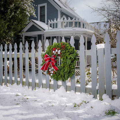 A holiday wreath made of noble fir and white pine with white jingle bells, 3 red berry clusters, 5 Australian pinecones, 2 red ball clusters, and a brushed red linen bow with white edging on a white picket fence.