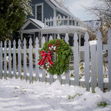 A holiday wreath made of noble fir and white pine with white jingle bells, 3 red berry clusters, 5 Australian pinecones, 2 red ball clusters, and a brushed red linen bow with white edging on a white picket fence.