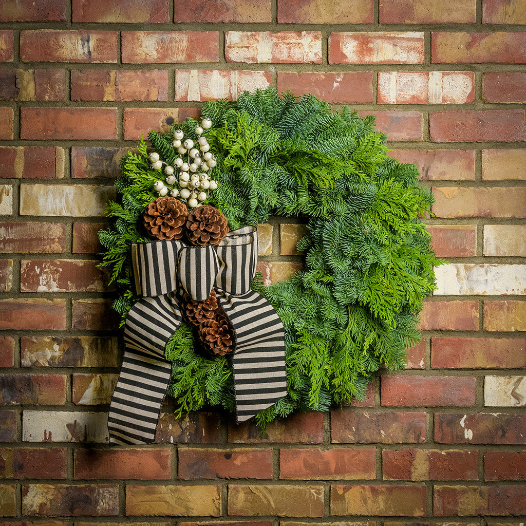 noble fir and cedar with 4 ponderosa pine cones, a branch of faux white berries, and a black and ivory striped burlap bow hanging on a brick wall