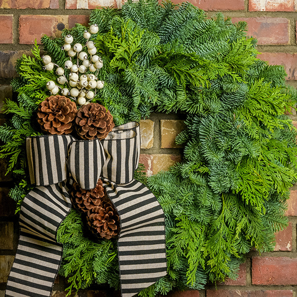 A wreath made of noble fir and cedar with 4 ponderosa pine cones, a branch of faux white berries, and a black and ivory striped burlap bow hanging on a brick wall.