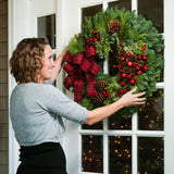 Christmas wreath made with pine cones, apples and berries and a red and black plaid bow being hung on a door by a woman