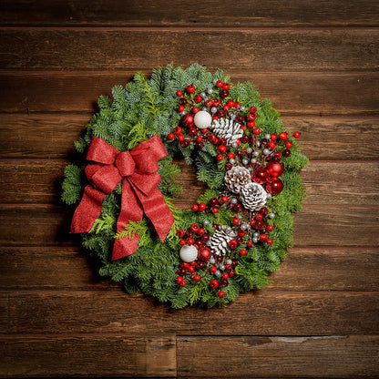 A wreath made of noble fir and Western red cedar with frosted ponderosa pinecones, red and white balls, red crabapples and silver berries, and a red glittery bow on a dark wood background.