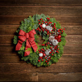 A wreath made of noble fir and Western red cedar with frosted ponderosa pinecones, red and white balls, red crabapples and silver berries, and a red glittery bow on a dark wood background.
