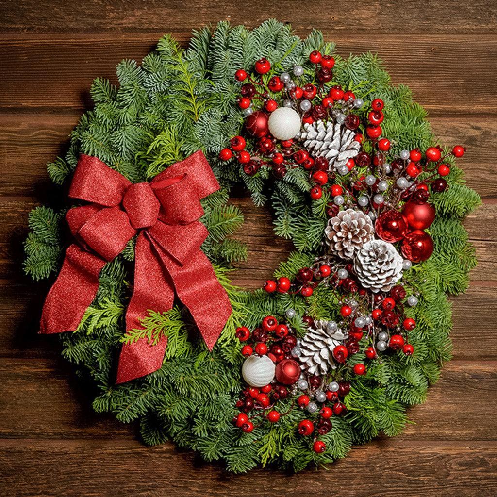 A wreath made of noble fir and Western red cedar with frosted ponderosa pinecones, red and white balls, red crabapples and silver berries, and a red glittery bow on a dark wood background.