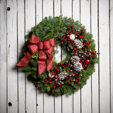 A wreath made of noble fir and Western red cedar with frosted ponderosa pinecones, red and white balls, red crabapples and silver berries, and a red glittery bow on a white wood background.