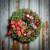 A wreath made of noble fir and Western red cedar with frosted ponderosa pinecones, red and white balls, red crabapples and silver berries, and a red glittery bow on a rustic metal background.