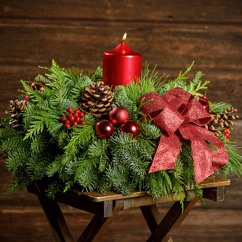 An arrangement made of noble fir, cedar, and white pine with natural pinecones, red berry clusters, red ball ornaments, a glittery red bow, and a metallic red pillar candle with a dark wood background.