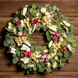 A dried wreath made of Preserved green silver dollar eucalyptus leaves, natural caspia, white globe amaranthus, natural and pink integrifolia, pink and white strawflowers on a dark wooden background.