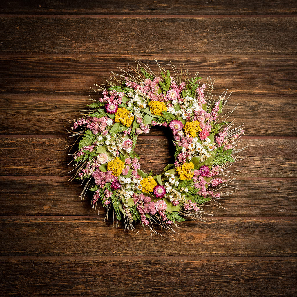 A dried wreath made of natural salal leaves, pink larkspur, natural ammobium, pink globe amaranthus, strawflowers, fern, and yellow coxcomb on a dark wood background.