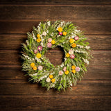 A wreath made of green caspia, lime green phalaris, natural oats, almond oats, yellow and pink strawflowers hung on a dark wooden background.