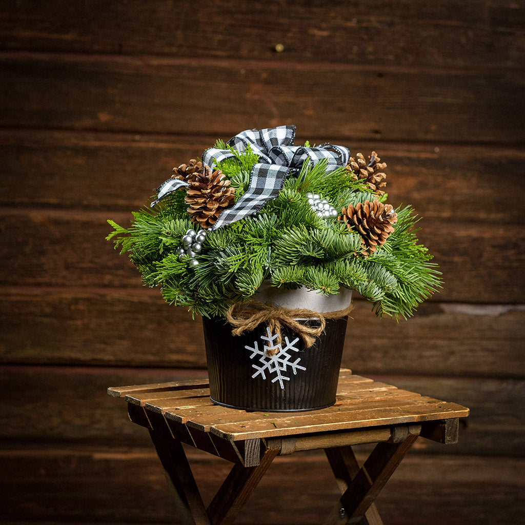 An arrangement made of noble fir, incense cedar, and white pine with Austrian pinecones, silver berry clusters, and a black and white plaid bow, black and white metal container, and white snowflake with a dark wooden background.