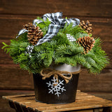 An arrangement made of noble fir, incense cedar, and white pine with Austrian pinecones, silver berry clusters, and a black and white plaid bow, black and white metal container, and white snowflake with a dark wooden background.