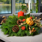  An arrangement made of white pine and salal with orange berry clusters, Austrian pinecones, festive sprays of autumn gourds, berries, seed pods, leaves, and an orange pillar candle sitting on a counter.