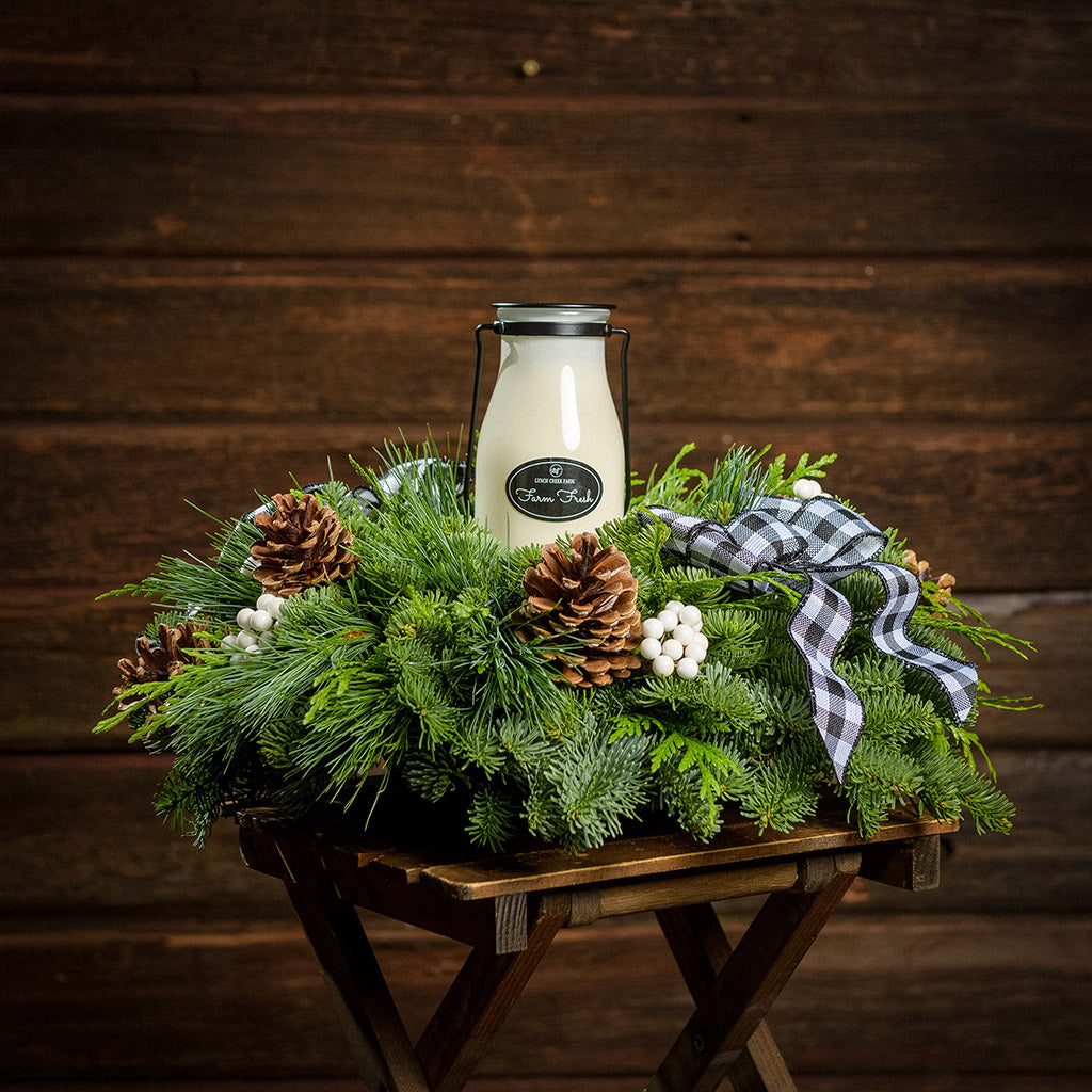 An arrangement made of noble fir, incense cedar, and white pine with Austrian pinecones, white berry clusters, a black and white gingham linen bow, and a milk bottle candle with an evergreen fir scent with a dark wooden background.