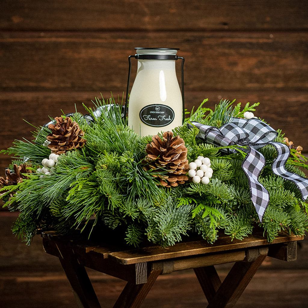 An arrangement made of noble fir, incense cedar, and white pine with Austrian pinecones, white berry clusters, a black and white gingham linen bow, and a milk bottle candle with an evergreen fir scent with a dark wooden background.