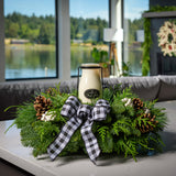 An arrangement made of noble fir, incense cedar, and white pine with Austrian pinecones, white berry clusters, a black and white gingham linen bow, and a milk bottle candle with an evergreen fir scent sitting on a counter.