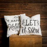 Set of 2 16 inch Happy Holidays and Let it Snow accent pillows in cream with black lettering on a wooden background. 