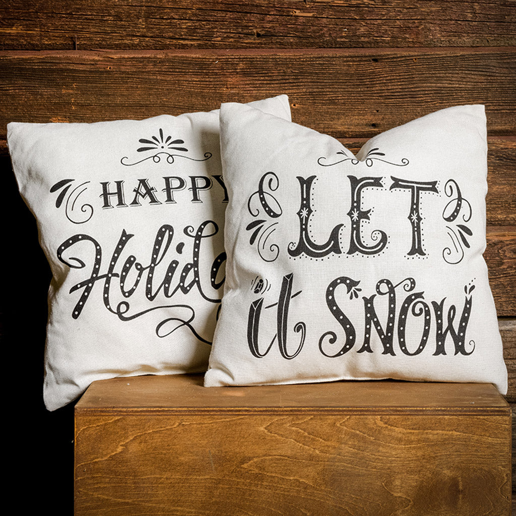 Set of 2 16 inch Happy Holidays and Let it Snow accent pillows in cream with black lettering on a wooden background.
