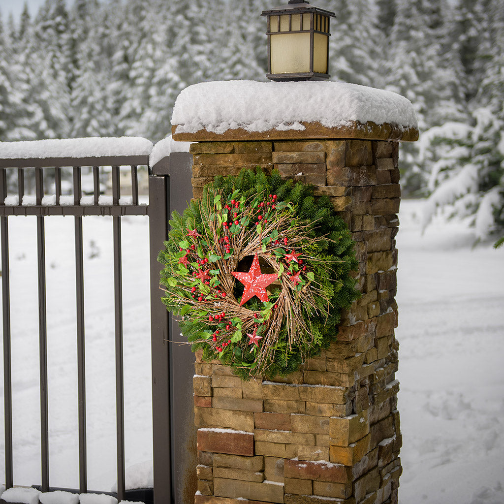 Farmhouse holiday Christmas wreath with speckled red, rustic stars, berries, holly and twigs on a base of noble and cedar on a natural brick pillar.