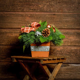 An arrangement made of White pine and salal with copper-orange balls, silver berry clusters, Austrian pinecones, and an orange plaid linen bow in an orange wooden container on a dark wood background.