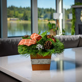 An arrangement made of White pine and salal with copper-orange balls, silver berry clusters, Austrian pinecones, and an orange plaid linen bow in an orange wooden container on a counter.
