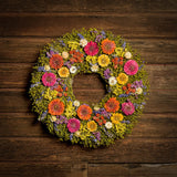 A dried wreath made of dried yellow, pink, and orange zinnias, lavender larkspur blooms, yellow statice flowers, and white strawflowers on a base of green flax podson hung on a dark wood background.